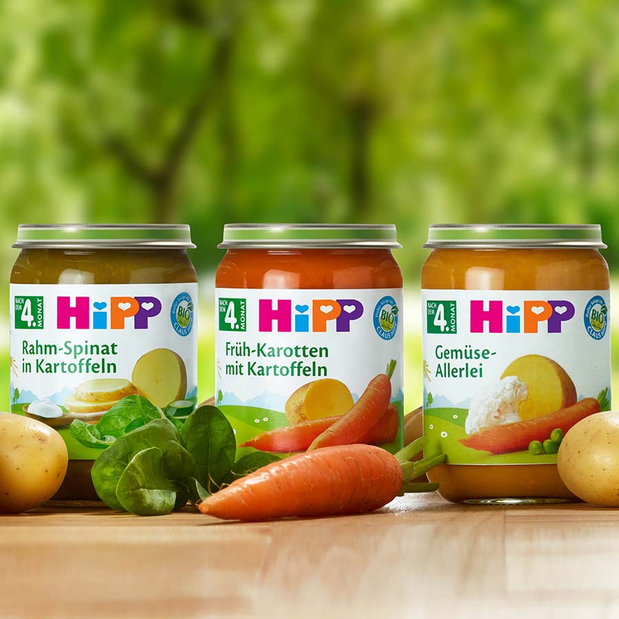 Three jars of baby food with labels in German are displayed on a wooden surface, surrounded by raw vegetables.